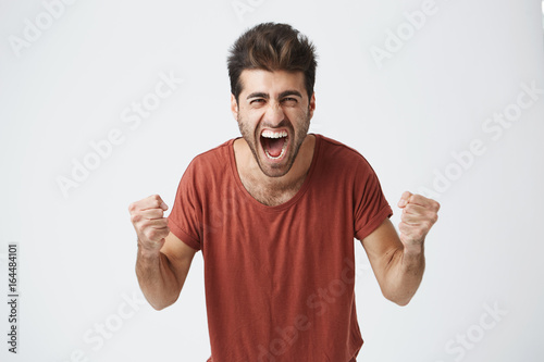 Happy positive excited young man clenching fists and screaming, wearing casual t-shirt glad to hear good news, celebrating his victory or success. Life achievement, goals and happiness concept.