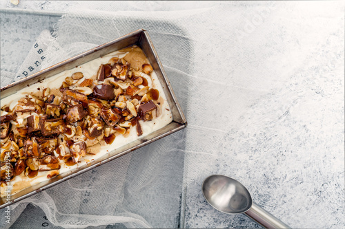 Snickers ice cream with caramel,  fried peanuts and hazelnuts in metallic baking tray on rustic background. Gourmet summer treat concept. Composition with copy space. photo
