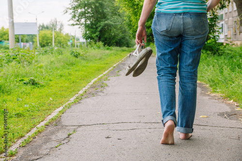 A woman walks barefoot along the asphalt road and holds shoes in her hands. Natural, Freedom
