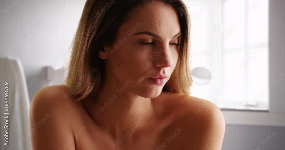 Close up of fresh faced young woman looking off camera with serious expression in bathroom. Casual portrait of attractive female millennial with bare shoulders sitting by window in bathroom. 4k Close up of fresh faced young woman looking off camera with serious expression in bathroom. Casual portrait of attractive female millennial with bare shoulders sitting by window in bathroom. 4k