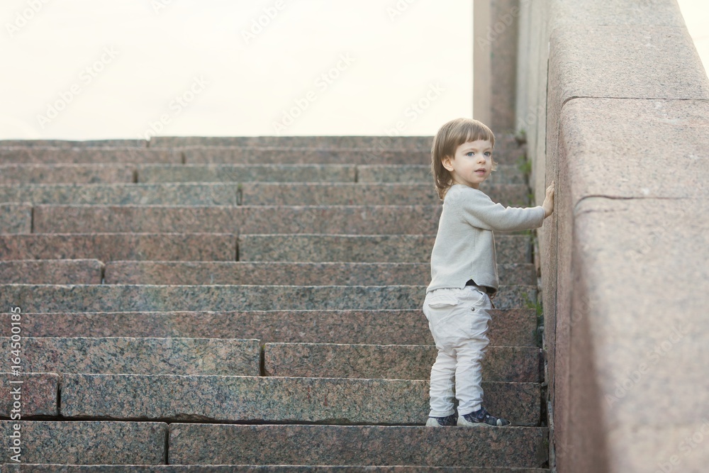 Little blue-eyed boy with long hair standing in front of the stone stairs. The concept of growing up