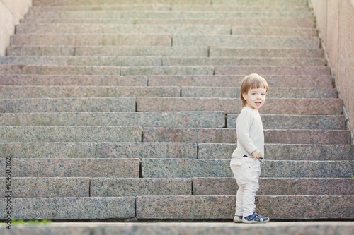 Little blue-eyed boy with long hair standing in front of the stone stairs. The concept of growing up