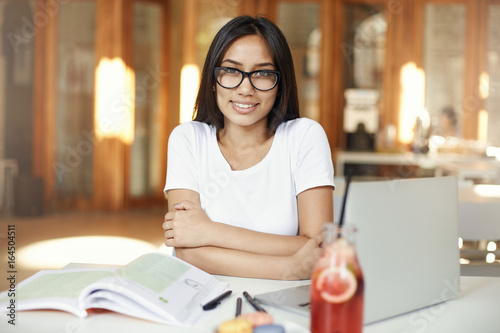 Portrait of assian woman smiling working on laptop in cafe or bright coworking space wearing glasses. photo