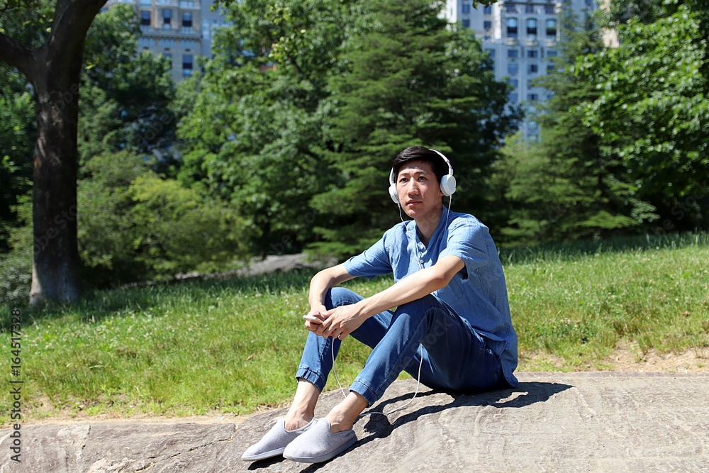 Young asian man sitting in urban city park outside listening to music with white headphones while holding smartphone Young asian man sitting in urban city park outside listening to music with white headphones while holding smartphone