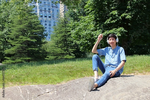 Young asian man sitting outside in city urban park listening to music with headphones while holding cell phone photo