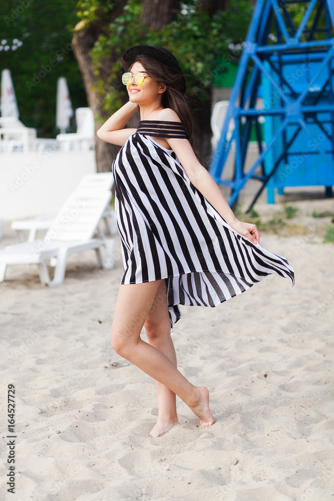 Luxury travel woman in black and white beachwear walking taking a stroll on sand summer beach. Girl tourist on summer holiday holding sun hat and yellow sunglasses at vacation resort Luxury travel woman in black and white beachwear walking taking a stroll on sand summer beach. Girl tourist on summer holiday holding sun hat and yellow sunglasses at vacation resort