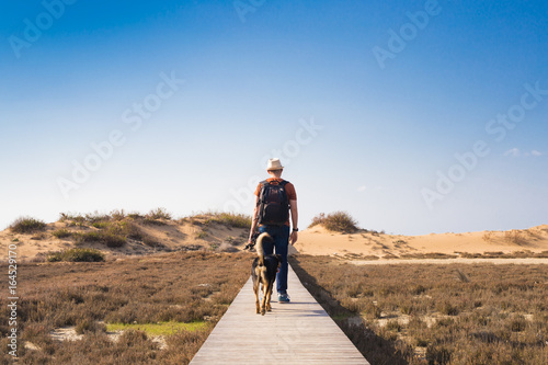 View from behind of a man walking with his dog on a road leading through beautiful landscape
