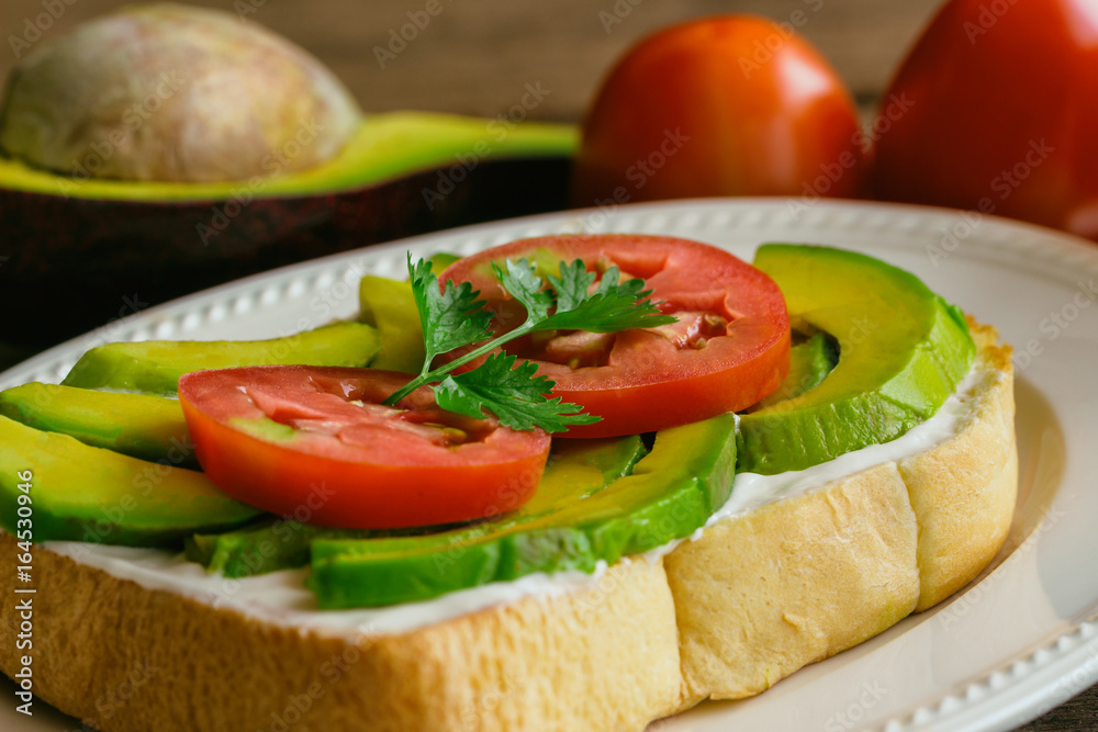 Open sandwich style for breakfast or lunch. Sandwich spread with cream cheese, avocado, tomato and parsley on white plate. Avocado open sandwich style serve with orange juice on rustic wood table.