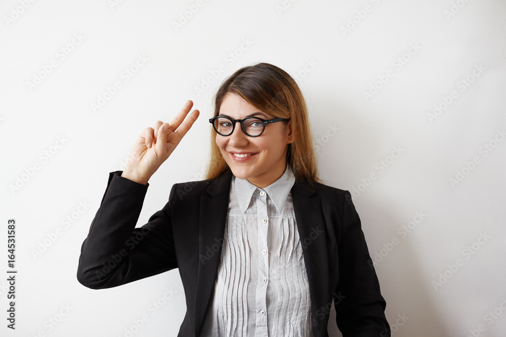 Indoor shot of successful and attractive young businesswoman wearing eyeglasses and formal jacket smiling broadly, showing victory sign at camera, posing against white studio wall background Indoor shot of successful and attractive young businesswoman wearing eyeglasses and formal jacket smiling broadly, showing victory sign at camera, posing against white studio wall background