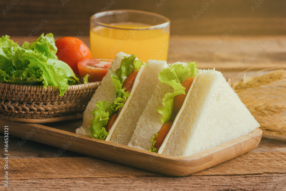 Piece of sandwich ham cheese with lettuce and tomato on wood plate. Homemade sandwich served with orange juice for breakfast or lunch. Delicious ham and cheese sandwich ready to served on wood table. Piece of sandwich ham cheese with lettuce and tomato on wood plate. Homemade sandwich served with orange juice for breakfast or lunch. Delicious ham and cheese sandwich ready to served on wood table.