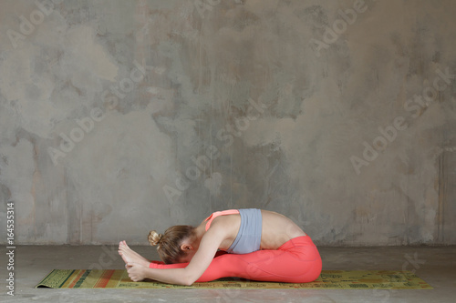 Young woman practicing left seated forward bend / Paschimottanasana yoga pose against texturized wall / urban background photo