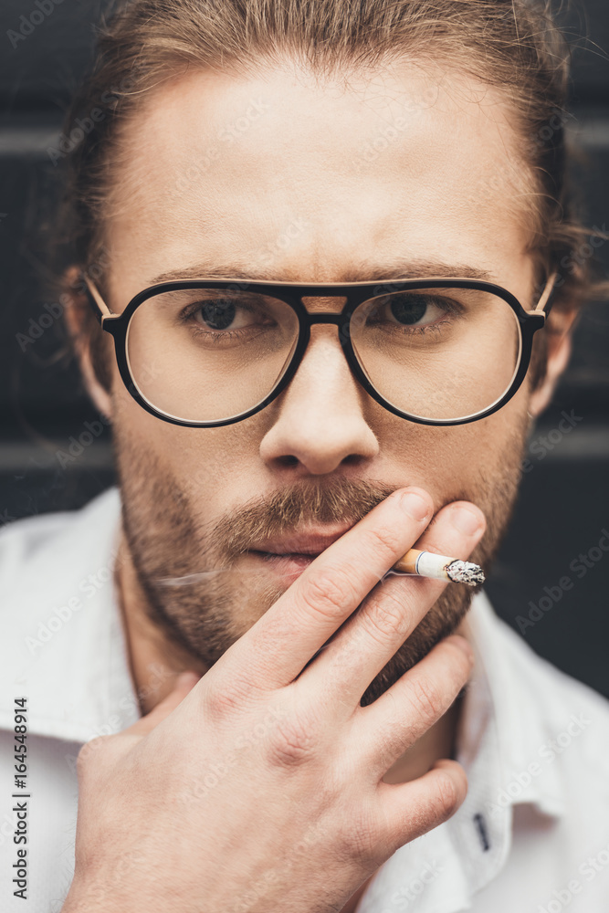 Close-up portrait of handsome stylish young man in eyeglasses smoking cigarette
