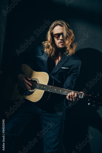 Handsome stylish man in suit and sunglasses playing guitar on black
