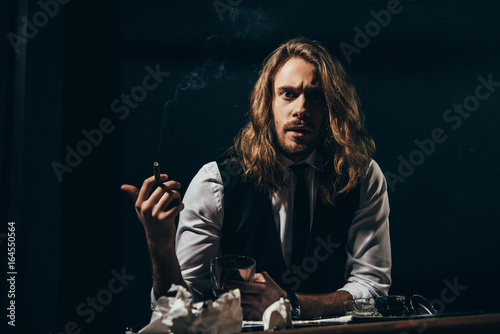 Bearded handsome long haired man in formal wear smoking cigar while holding glass of whisky and looking at camera