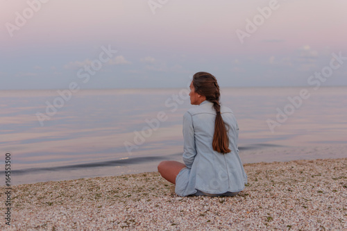 Young beautiful lonely girl is sitting on the beach and looking into the distance of the sea at sunset (dawn). 