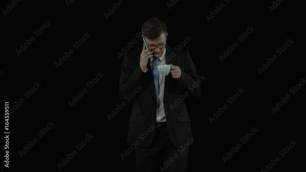 Handsome businesman walks over black backgroung speaking on the phone and drinking coffee from a cup Handsome businesman walks over black backgroung speaking on the phone and drinking coffee from a cup