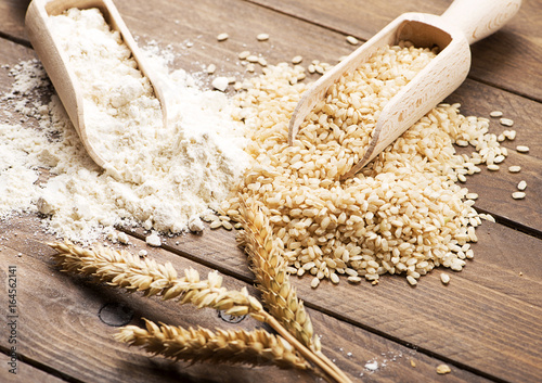 Wooden spoon with flour next to rice and wheat flower on brown wooden table.