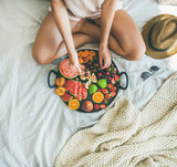 Summer healthy raw vegan clean eating breakfast in bed concept. Young girl wearing pastel colored home clothes taking cherries from tray full of fresh seasonal fruit. Top view, square crop, copy space