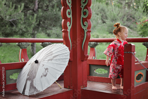 Small toddler redhead girl in traditional Japan National Costume yukata with oil-paper umbrella Wagasa or Bangasa on the background of Asian alcove. multicultural society concept photo