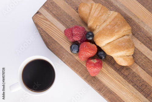 Croissant with blueberries and raspberries on a chopping board with black coffee