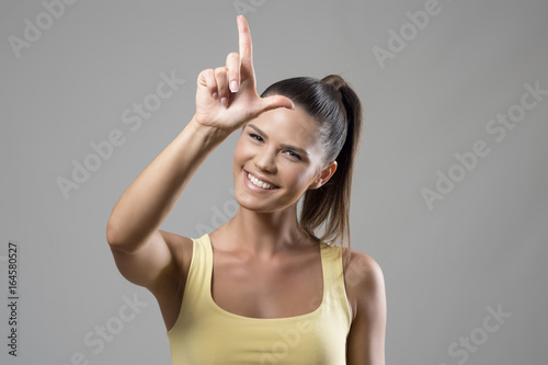 Photography Young fit sporty woman showing letter L loser hand gesture on forehead smiling at camera over gray studio background