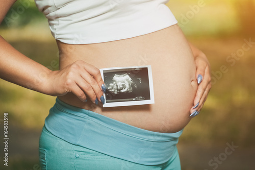 Photos Adult pregnant woman holding a photo Medical ultrasound near the abdomen on a nature background