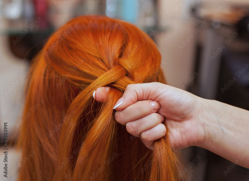 Fototapeta premium Beautiful girl with red long hair, hairdresser weaves a French braid, in a beauty salon. Professional hair care and creating hairstyles.
