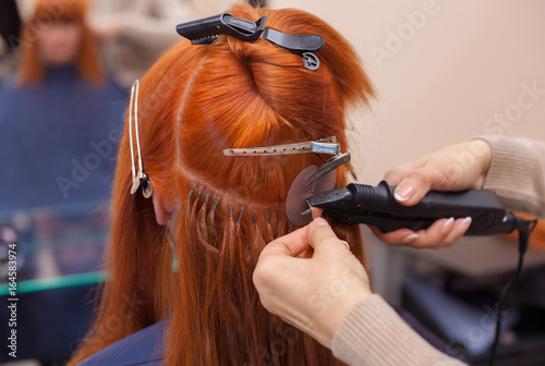 The hairdresser does hair extensions to a young, red-haired girl, in a beauty salon. Professional hair care.