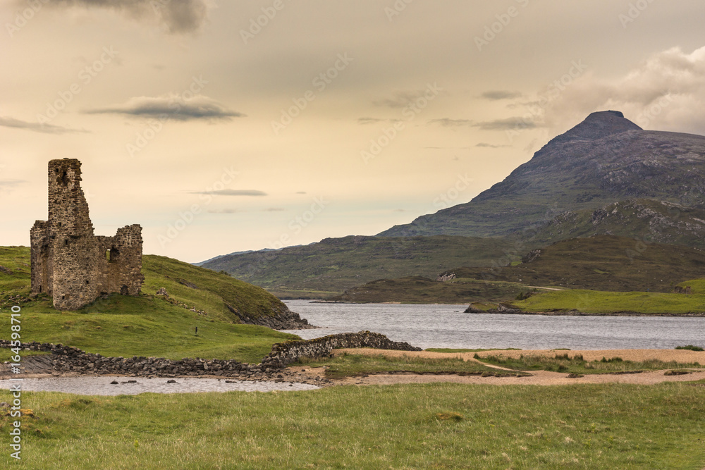 Assynt Peninsula, Scotland - June 7, 2012: Ruins of Castle Ardvreck at Loch Assynt with mountain in back under yellowish twilight sky. Green ground vegetation.