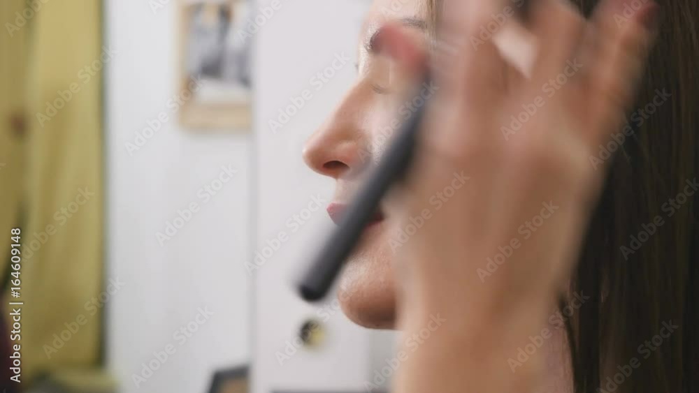 Visagiste corrects a young woman's face with broad make-up brush