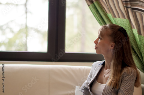 Portrait lady with long hair in a grey suit sits thoughtfully at the bright window.