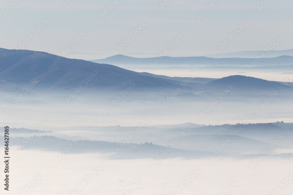 A view from above of a valley filled by a sea of fog, with various layers of emerging hills and mountains with different shades of blue