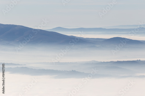 A view from above of a valley filled by a sea of fog, with various layers of emerging hills and mountains with different shades of blue