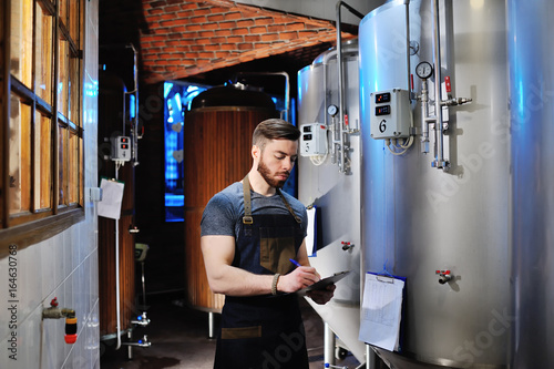 A handsome bearded male brewer records data against a background of beer tanks. Brewing