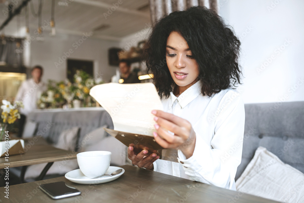 Beautiful girl in white shirt sitting in restaurant with menu in hands. Young lady with dark curly hair thoughtfully looking in menu at cafe. African American girl with cup on table in cafe Beautiful girl in white shirt sitting in restaurant with menu in hands. Young lady with dark curly hair thoughtfully looking in menu at cafe. African American girl with cup on table in cafe