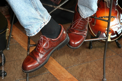 Musician wearing shiny brown shoes, faded blue jeans and a guitar close by.