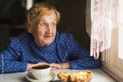 An old woman sitting near the window drinking tea with buns.
