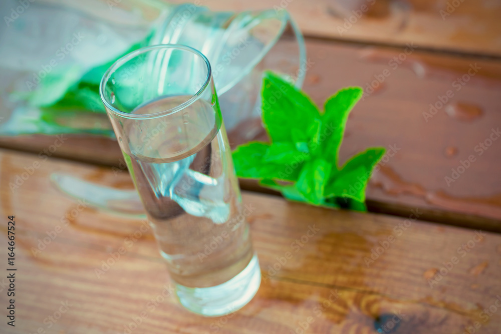 A glass with fresh water next to a jug with mint on a wooden table close up blurred A glass with fresh water next to a jug with mint on a wooden table close up blurred
