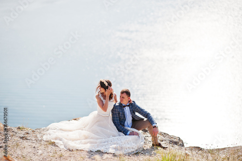 Stunning young wedding couple sitting on the edge of the cliff with a beautiful scenery on the background. © AS Photo Family