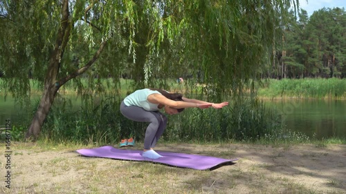 Flexible flexible young attractive girl doing exercises from yoga. A woman is sitting on a foil rug on the lake's bregue and performing the dove posture. Asana of awakening. Improvement of blood