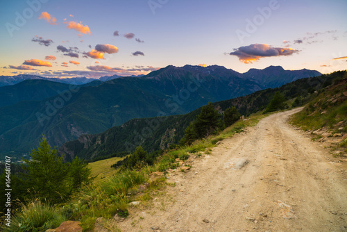Dirt mountain road leading to high mountain pass in Italy. Expasive view at sunset, colorful dramatic sky, adventures in summer time, Italian Alps.