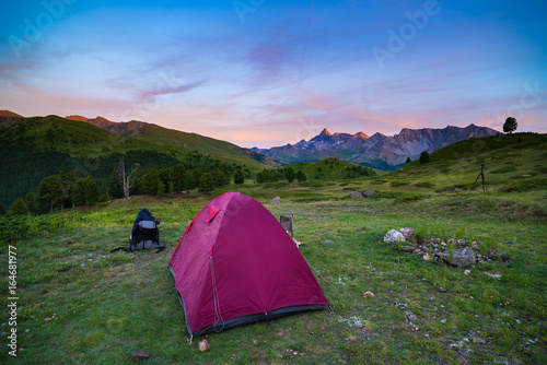 Camping with tent at high altitude on the Alps. Snowcapped mountain range and scenic colorful sky at sunset. Adventure and exploration in summertime.