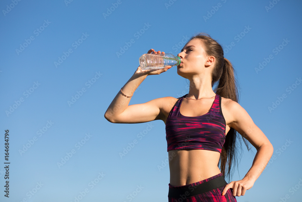 Young happy woman drinking water from bottle. Isolated sportswoman in sportswear holding drink outdoors at blue sky background. She quenches thirst. Young happy woman drinking water from bottle. Isolated sportswoman in sportswear holding drink outdoors at blue sky background. She quenches thirst.