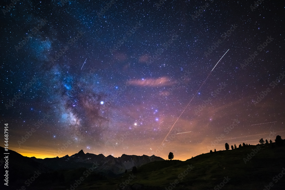 Fototapeta premium The colorful glowing core of the Milky Way and the starry sky captured at high altitude in summertime on the Italian Alps, Torino Province. Airplane traffic in the sky.