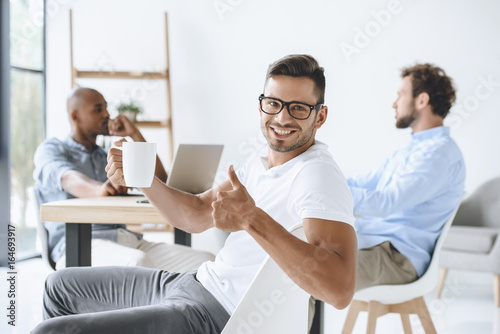 smiling businessman with cup of coffee showing thumb up in office