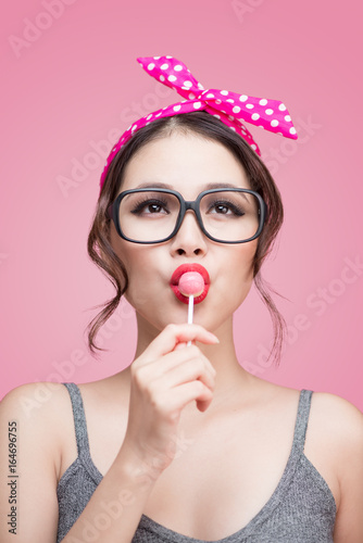 Portrait of beautiful asian woman eating heart shape lollipop, dressed and makeup in pin-up style on pink background.