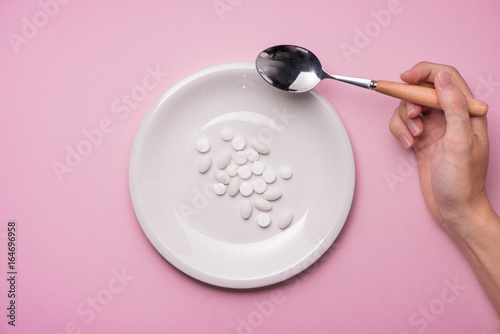 Top view of man's hands at dining table holding a fork and knife above dish with pills over pink background.