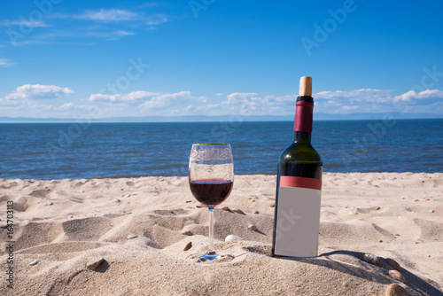 A glass of red wine and a bottle on the beach in a summer sunny day. Sea and blue sky in the background
