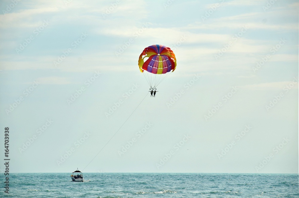 Beach vacation at sea. Holidaymakers tourists fly high in sky on multi-colored parachute, which pulls boat on its rope Beach vacation at sea. Holidaymakers tourists fly high in sky on multi-colored parachute, which pulls boat on its rope