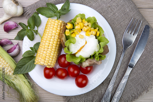 The poached egg in the basket of bacon with herbs and tomatoes and corn with garlic. On wooden background.